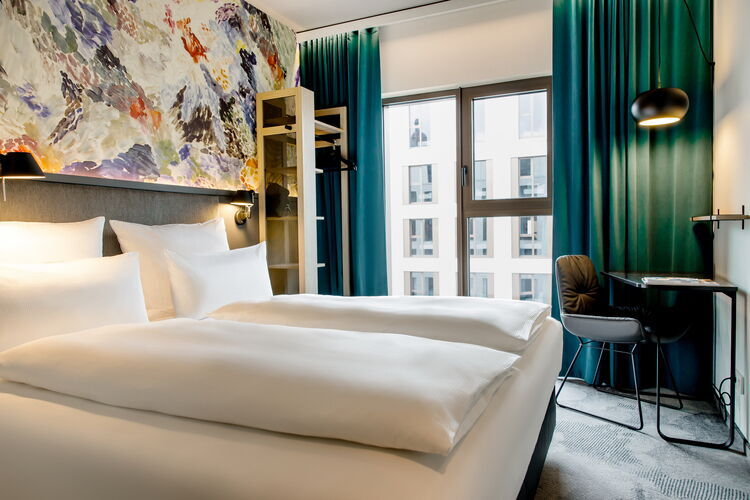 Modern hotel room with two white box-spring beds, colourful wall decorations and large windows with turquoise curtains at Motel One Antwerp.