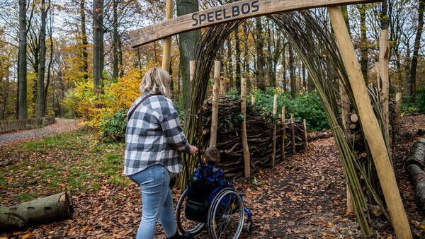 Een vrouw wandelt met een kindje in de rolstoel door een speelbos