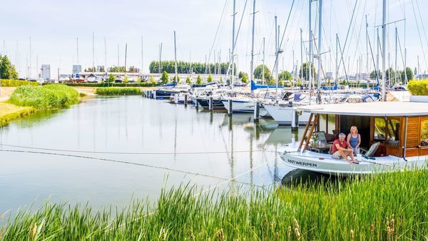 A man and a woman on a sailboat in green surroundings. In the background, other sailboats.