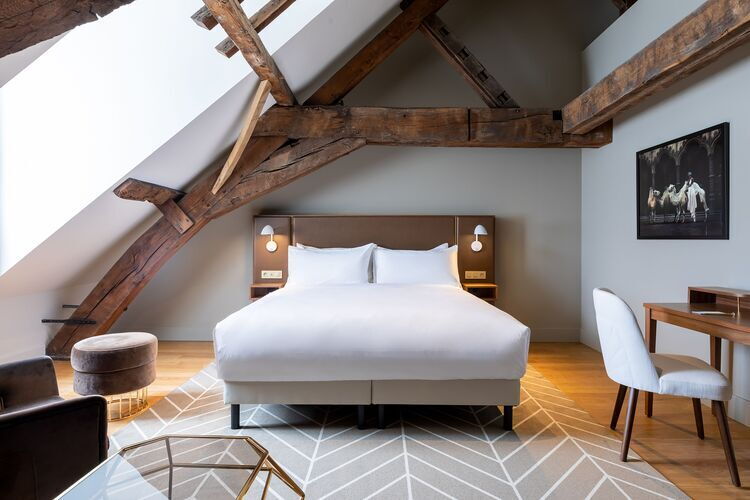 Stijlvolle hotelkamer onder een houten dakgebinte, met een groot tweepersoonsbed, minimalistische nachtlampen, een zacht vloerkleed en een zithoek met bureau en kunstwerk aan de muur.