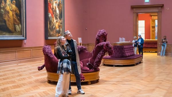 Une femme et un homme regardent un tableau dans une salle majestueuse du Musée royal des Beaux-Arts d'Anvers. En arrière-plan, des personnes se promènent et d'autres peintures sont accrochées au mur.