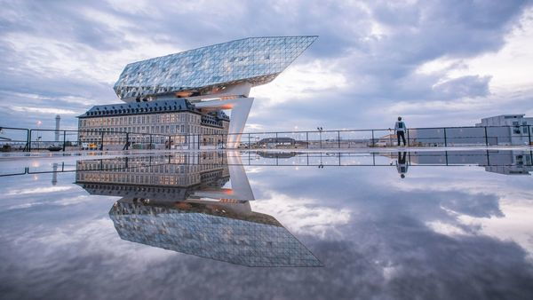 A classical building with a modern building above it in the shape of a diamond ship reflects in the water. A man looks at the building.