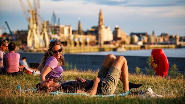 A man and a woman are laughing on the grass along the Scheldt. In the background, the skyline of the Historical Centre including the cathedral.