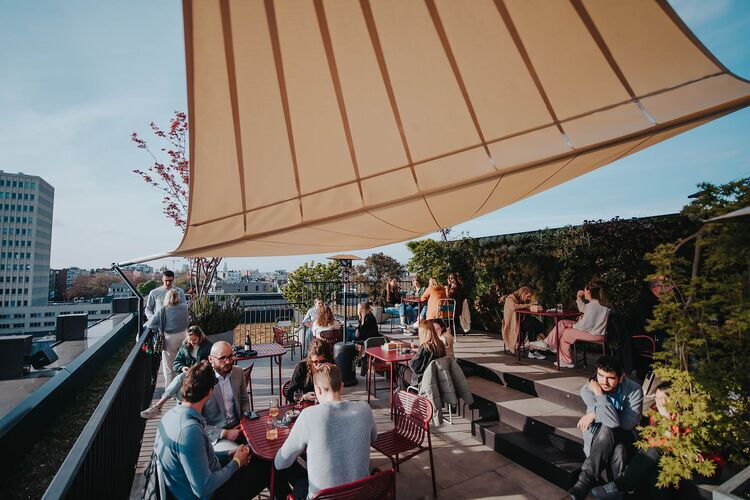Dakterras Yust Antwerp met mensen die ontspannen aan tafels zitten onder een groot zonnedoek, omringd door groen en met uitzicht over de stad.