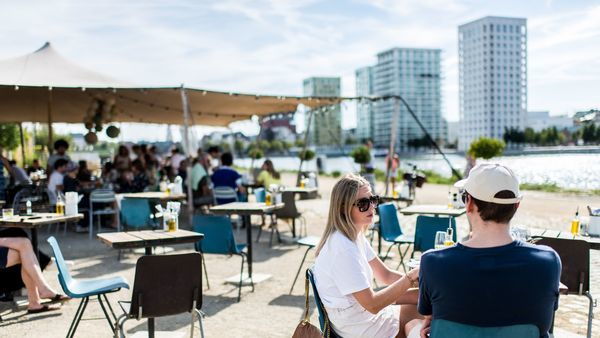 A man and a woman sit on a waterfront terrace. In the background, more people are sitting and some skyscrapers.