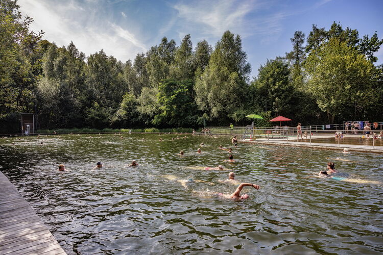 Des visiteurs se détendent au bord de l'étang de baignade Boekenberg à Anvers, avec de l'eau naturelle, des pontons en bois et de la verdure tout autour.