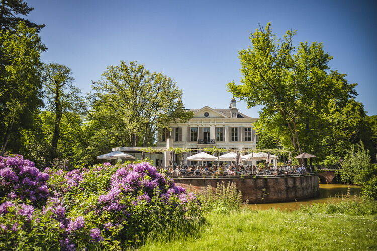 Historisch landhuis met groot tuin­terras aan het water, omringd door groene bomen en bloeiende rododendrons in een landschapspark.