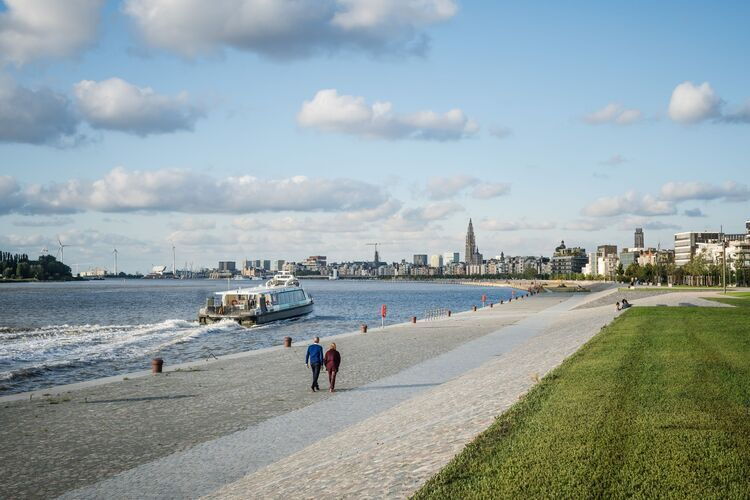 Zwei Personen spazieren entlang der renovierten Uferpromenade der Schelde in Antwerpen, mit Blick auf die Schelde, ein vorbeifahrendes Schiff und die Skyline von Antwerpen im Hintergrund.