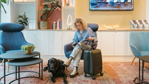 Dog lies on floor at hotel lobby, owner in seat browsing magazine.