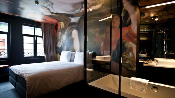 Una elegante habitación de hotel con cama y baño. Las paredes y el techo están pintados o empapelados con figuras y escenas rubensianas.
