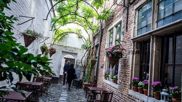 A few people stroll down a quaint and medieval-looking street where terrace tables and chairs stand against the facades.