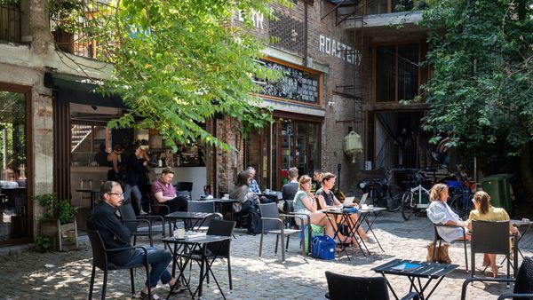 Several people sit at tables the outdoor terrace of a coffee shop in a green setting.