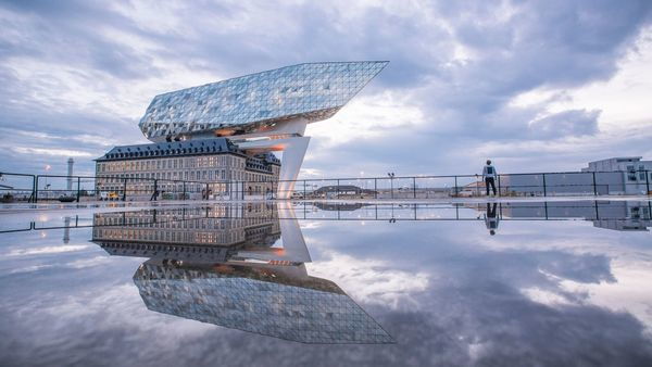 Atop a large 19th-century building is a modern superstructure in the shape of a diamond ship. The building reflects in the water; a man stands in the background.
