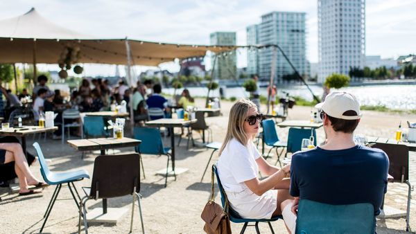 A man and a woman sitting on a terrace by the water. In the background, more people are sitting and there are some skyscrapers.