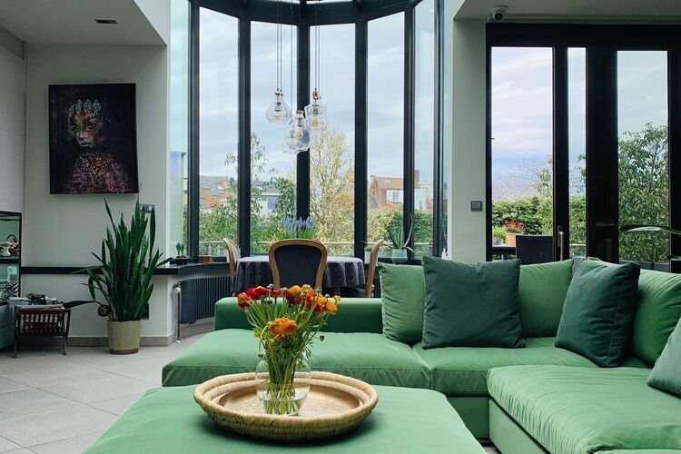 Bright living room at B&B Puur with large windows, a green corner sofa and a coffee table with a vase of fresh flowers, overlooking the garden and dining table in the background.