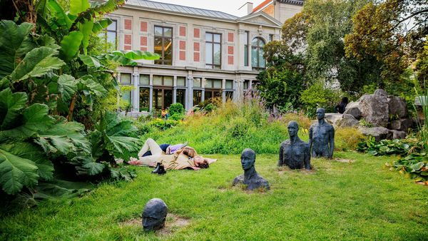 People lie on the grass in the Botanical Garden, a row of busts in the foreground and a classical building in the background.
