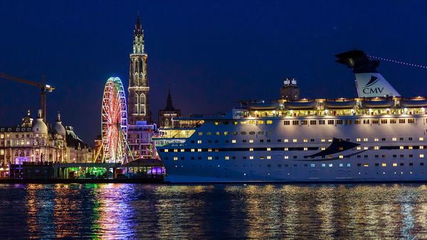 Cruise ship Magellan sails down the Scheldt after dark past the illuminated cathedral and a Ferris wheel. 