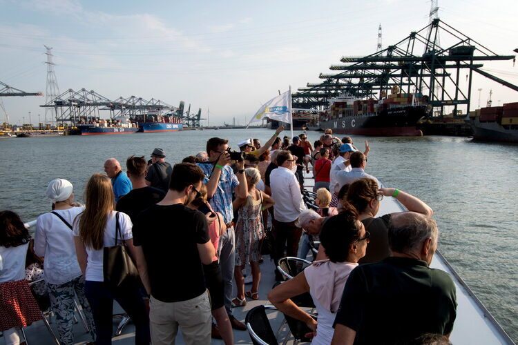 Passagiere stehen auf dem offenen Deck eines Schiffes auf der Schelde in Antwerpen, im Hintergrund sind Hafenkräne, Containerschiffe und die Hafenanlagen entlang der Uferpromenade zu sehen.