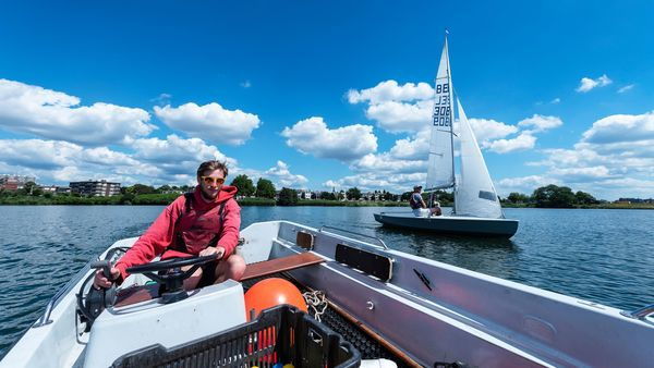A man at the wheel of a sailing boat. Next to him, another sailboat.