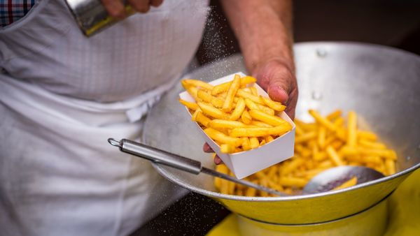 De handen van een man gieten zout over een bakje frieten. Het bakje wordt vastgehouden boven een frituurvergiet