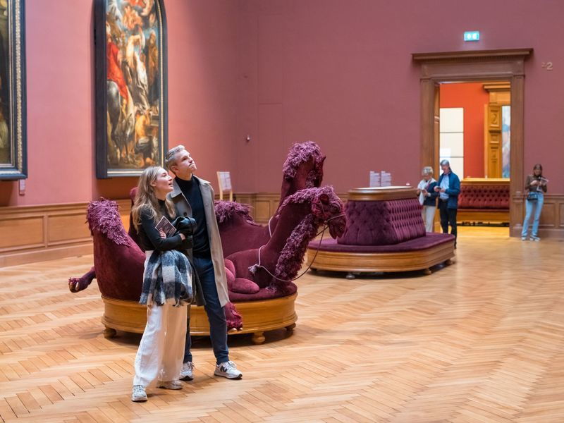 A woman and a man look at a painting in a stately museum room at the Royal Museum of Fine Arts Antwerp. In the background, some people are walking around and more paintings hang on the wall.
