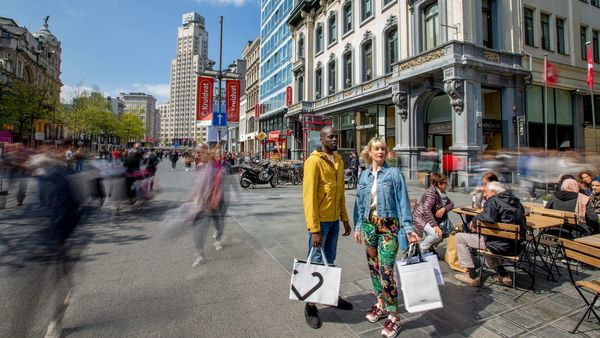 A man and a woman stand with shopping bags on the Meir. In the background, people walk past shops and the skyscraper Boerentoren stands.