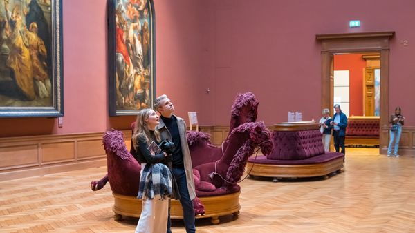 Een vrouw en een man kijken in een statige museumzaal van het Koninklijk Museum voor Schone Kunsten Antwerpen naar een schilderij. Op de achtergrond wandelen enkele mensen rond en hangen nog meer schilderijen aan de muur.