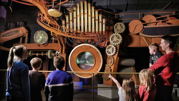 Several children and a man holding a small child are looking at a large 19th century machine filled with radars and air hoses.