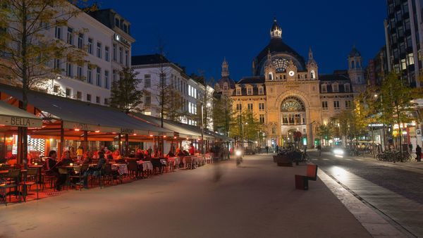 People sitting on terraces at night on De Keyserlei, in the background the façade of Central Station.