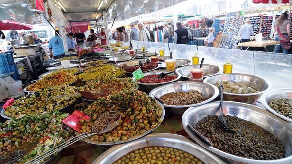 Des épices et des ingrédients exotiques sont exposés sur une table dans un marché. En arrière-plan, des gens passent.