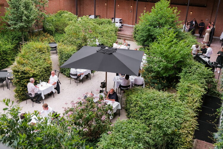 Green courtyard garden of Hotel August in Antwerp with an attractive terrace, surrounded by lush vegetation, seating with parasols and guests enjoying a peaceful outdoor dining experience.