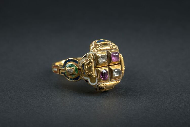 Close-up of a gold ring with a geometric design and coloured gemstones set against a dark background.