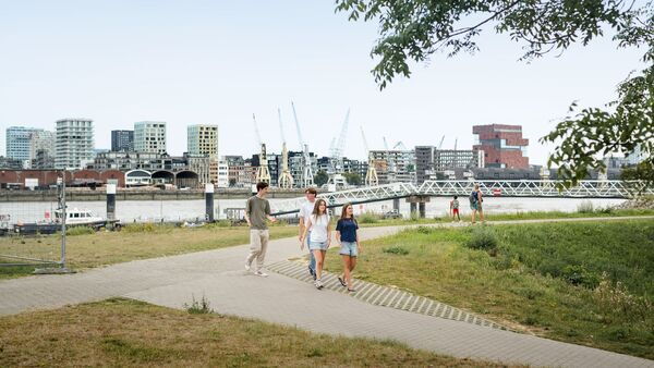 Spaziergänger auf einem Weg entlang der Schelde in Antwerpen, im Vordergrund Rasenflächen und im Hintergrund die Kräne der Stadt und des Hafens.