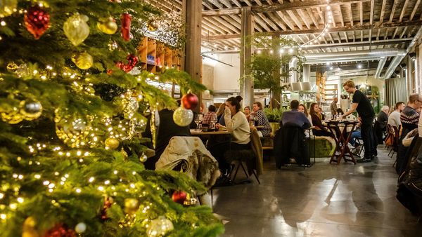 Menschen sitzen an einem Tisch und essen in einer Winterbar. Im Vordergrund steht ein Weihnachtsbaum.