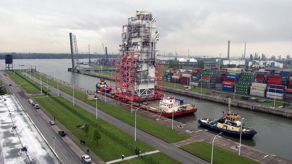In a harbour lock, a tugboat tows a pontoon on which a cooling tower stands. In the background stacked containers, the Scheldt and industry. 