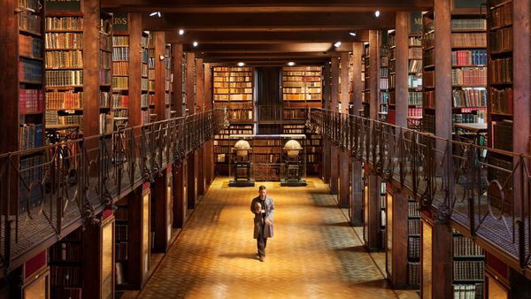Een imposante historische bibliotheekzaal met een hoog plafond. Op twee zijverdiepen staan kasten vol boeken.