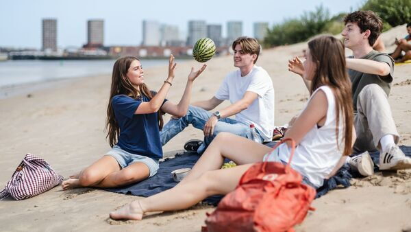 Einige Jugendliche sitzen gemeinsam am Strand von Sint-Anneke in Antwerpen und spielen mit einem Ball, im Hintergrund sind die Schelde und die Skyline von Antwerpen zu sehen.