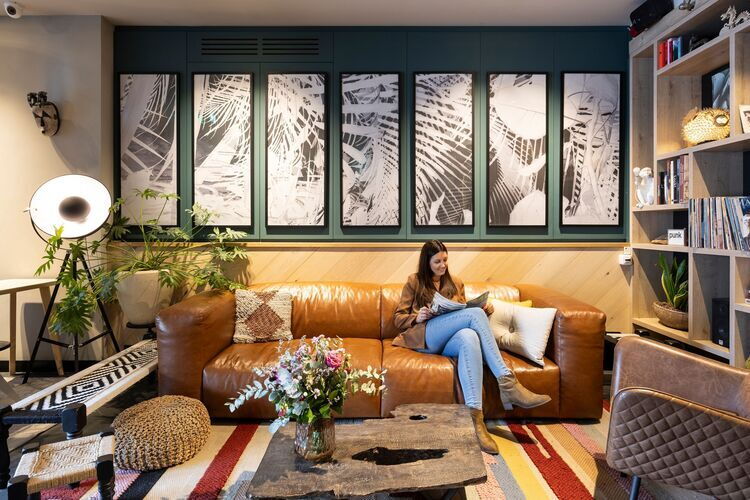 Cosy lounge area in Hotel Indigo in Antwerp, with a warm leather sofa, art on the walls, lots of plants and a guest relaxing and reading a magazine.