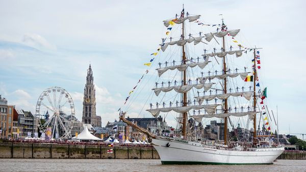 Dozens of sailors stand on the masts of their tall ship that is sailing on the Scheldt past the cathedral and the ferris wheel.