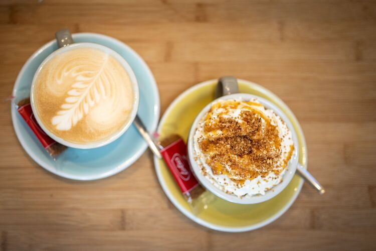 Twee koffies op een tafel, het blauw gekleurd kopje heeft een melkschuimpje in de vorm van een blad, het geel gekleurd kopje heeft een speciale topping. 