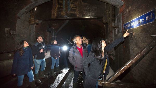 A group of people stand in a tunnel. A woman points to an old street name sign.