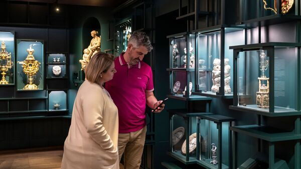 Koppel staat in een van de zalen van DIVA en kijkt naar gsm. Rond hen staan vitrines met zilveren objecten.. 