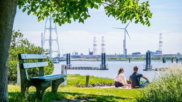 A woman and a man sit on a picnic blanket in green surroundings. In the background, the Scheldt River flows and there are windmills.